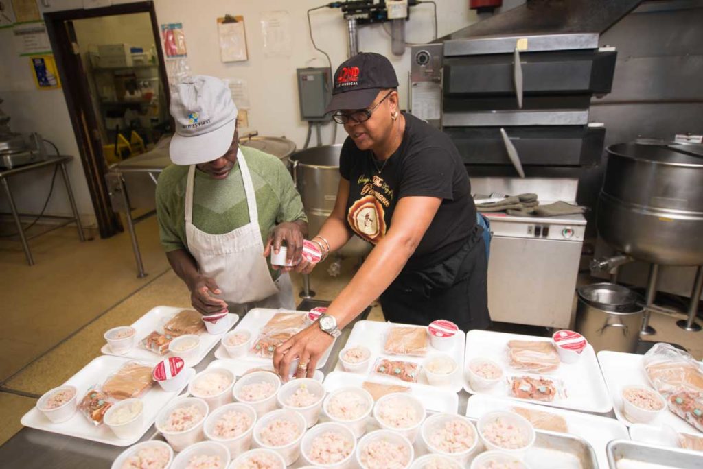 Two workers ration out food portions for cafeteria trays.