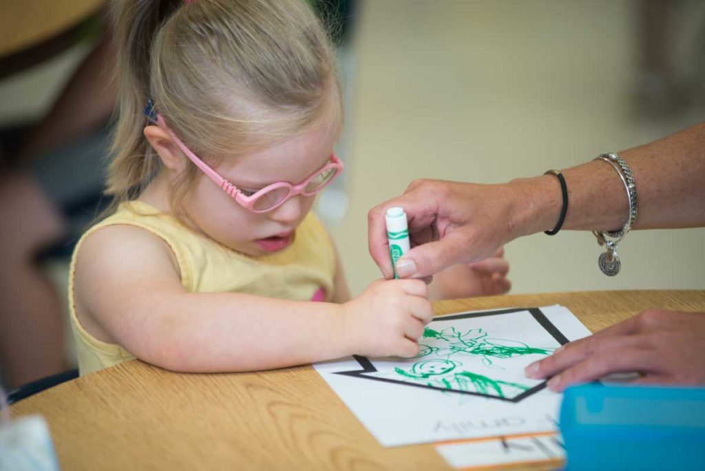 A woman helps a girl draw with a green marker.